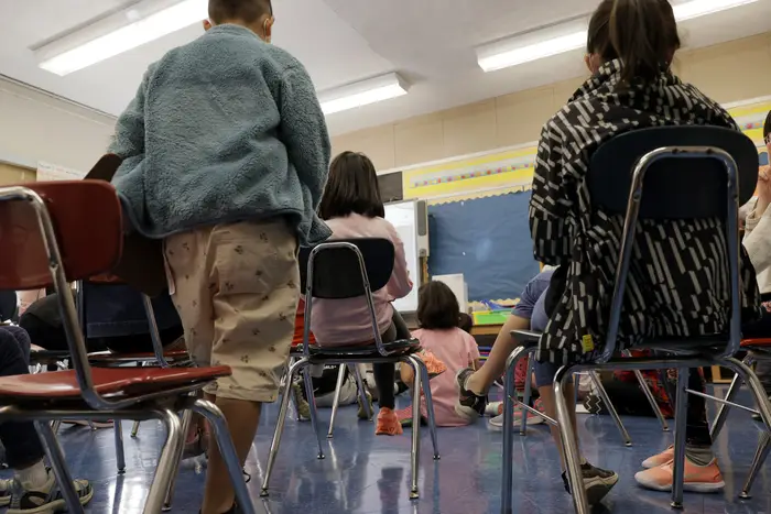 Inside a classroom, we see backs of students who are sitting in chairs and some who are sitting on the ground.